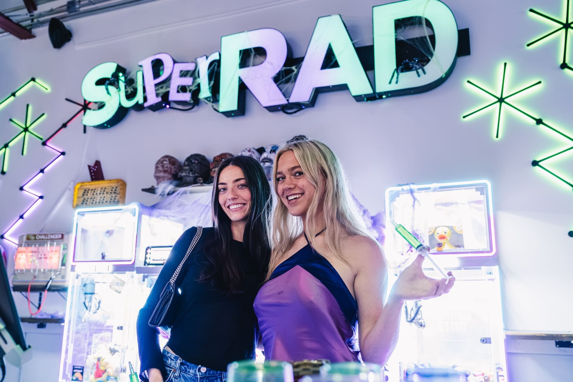 Two friends smiling under the Super Rad neon sign at the Raleigh arcade bar